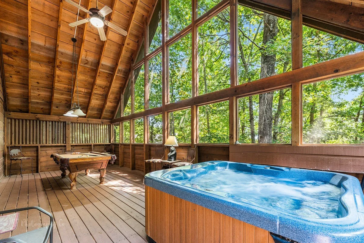 This image features a spacious interior entertainment area with a pool table and a hot tub, both set against large windows offering tranquil nature views. The mood is inviting and social, suggesting group activities like playing pool and relaxing in the hot tub while sunlight streams in or during cozy evenings.