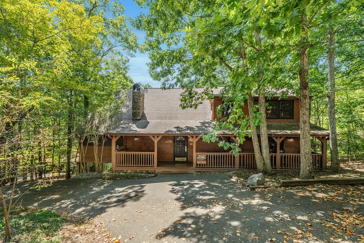 The image features a rustic wooden porch with a stone chimney, large front windows, and lush trees surrounding the cabin. The spacious porch is an inviting spot for morning coffee or evening relaxation, with nature all around.
