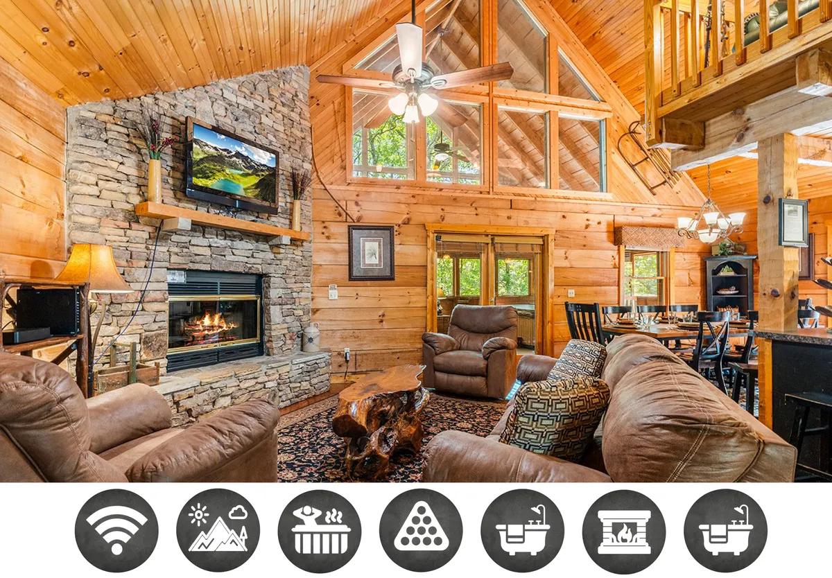 The image showcases a warm, wood-accented living space featuring a stunning stone fireplace as the centerpiece. Large windows frame the natural light and views, while the open-concept area seamlessly blends a cozy lounge with a spacious dining table, perfect for gatherings. The vaulted wooden ceiling and comfortable seating evoke a classic mountain cabin ambiance.