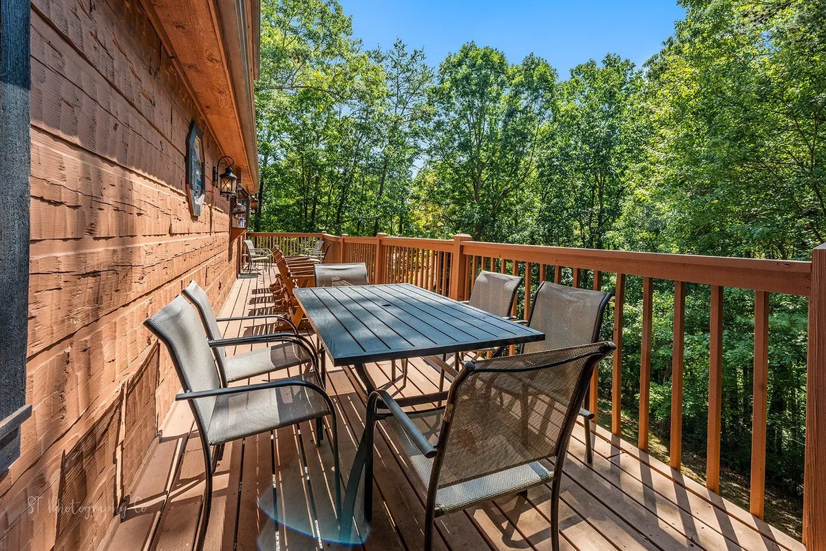 A wooden deck with a spacious outdoor dining table sits nestled among tall trees. The area is clearly set up for alfresco meals or gatherings, with seating that can accommodate family or friends. The deck's natural setting and open design embody the essence of mountain living.
