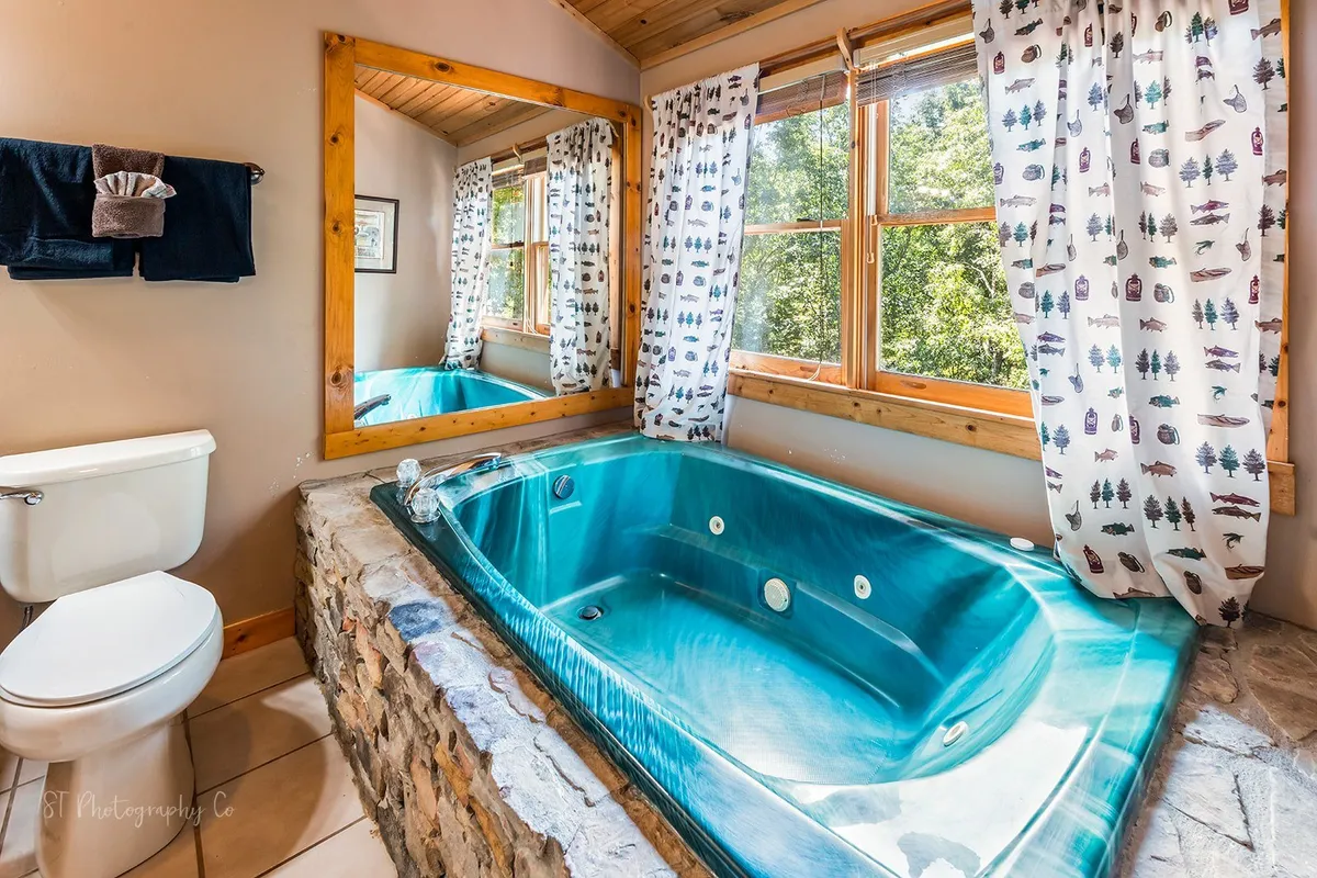 A luxurious bathroom with a jacuzzi tub positioned by large windows, a stone accent wall, wooden ceiling, and decorative curtains. Sunlight streams in, creating a spa-like ambiance ideal for relaxing soaks.