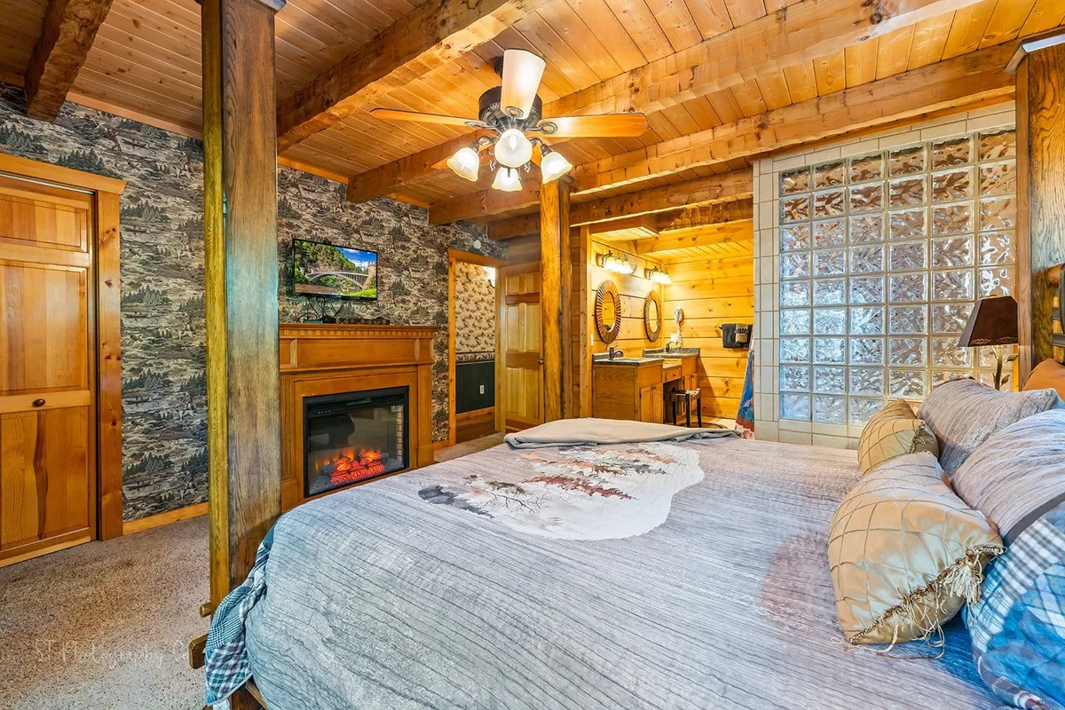 A cozy bedroom with a comfortable bed, wooden beams, a seasonal fireplace, decorative wallpaper, glass block wall, and soft lighting. The fireplace enhances the room's warmth and romantic mood.
