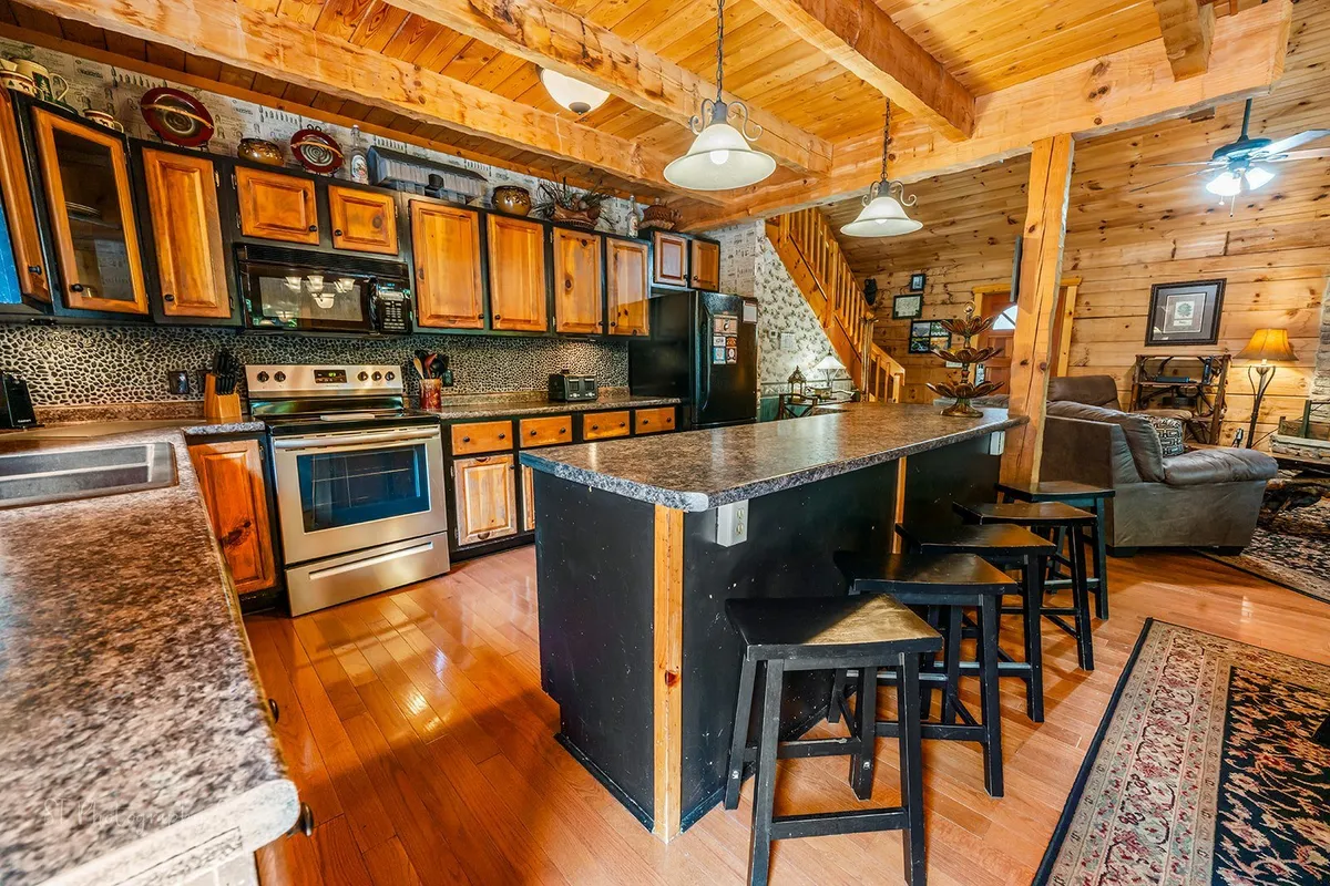 The image captures a view of the kitchen with an expansive island, wooden cabinetry, stainless steel appliances, a staircase in the background, and a comfortable seating area. The warm, inviting lighting and spacious layout invite guests to gather for meal prep and conversation.