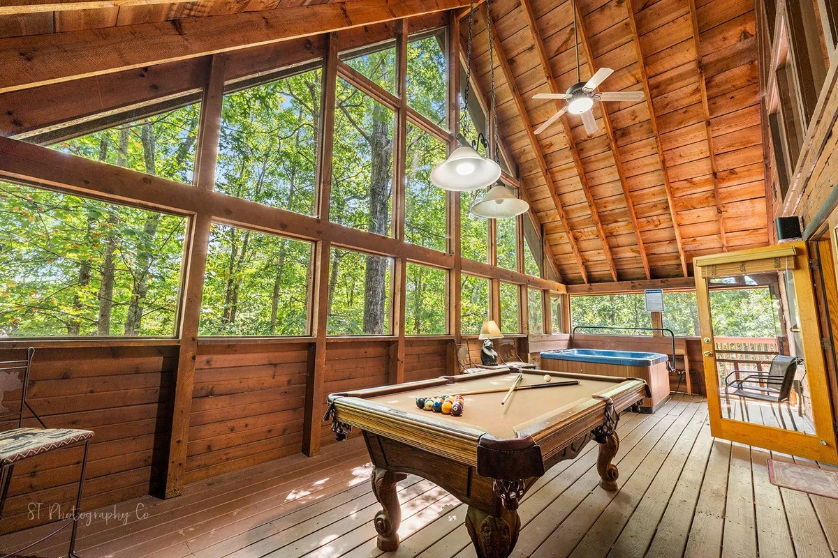 The image displays a light-filled entertainment area with a pool table and a hot tub, surrounded by large windows that frame beautiful views of the outdoors. The wooden ceiling and cozy layout set the scene for friendly competition and relaxation among friends.