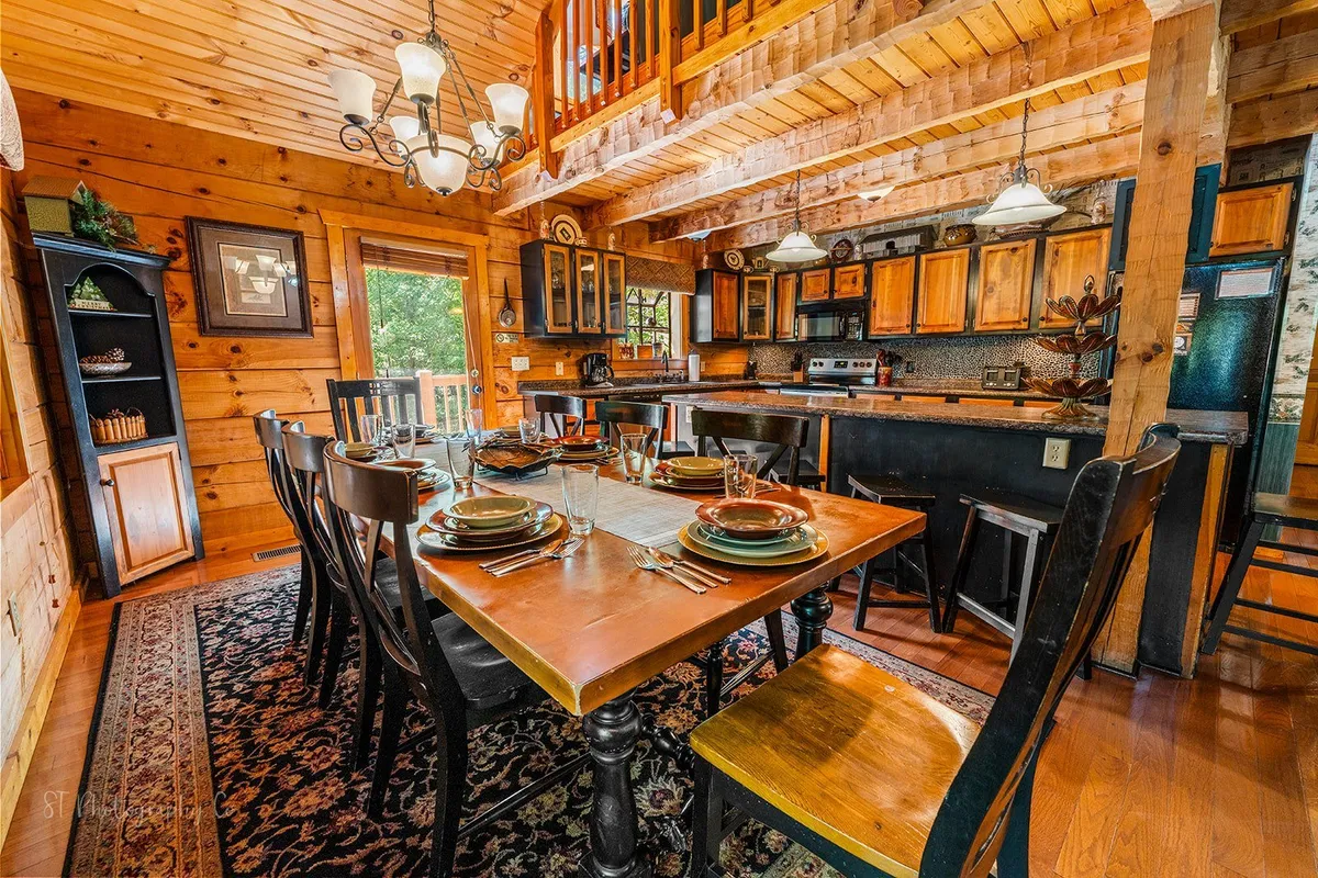 A beautiful dining area is set for a meal, featuring a wooden dining table with seating for eight or more, elegant chandelier lighting, and cozy rug. Wooden cabinetry in the background adds rustic warmth, creating an ideal setting for special gatherings.