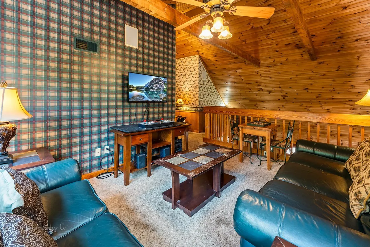 A cozy loft space with leather sofas, a coffee table, plaid wallpaper, and a TV creates an inviting setting for movie marathons, board games, and social gatherings. The warm lighting and comfortable furnishings set a playful and relaxing atmosphere.