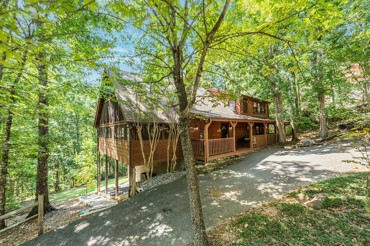 This exterior image shows the front of a cozy wooden cabin nestled among mature trees, with a driveway leading up to the entrance. The surrounding greenery hints at the cabin’s proximity to nature and local attractions, making it an ideal base for exploration.
