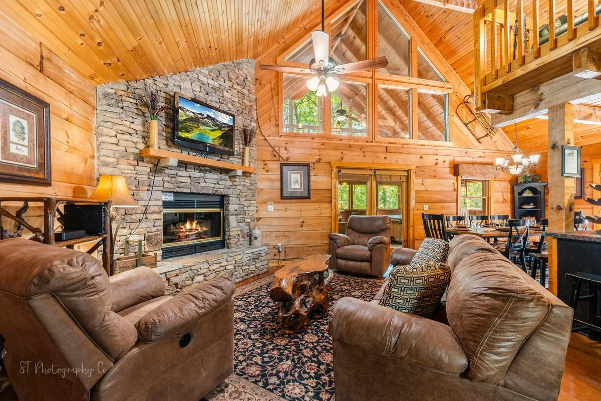 This image showcases a spacious living room with a prominent stone fireplace, large windows letting in natural light, and comfortable seating arranged around a decorative rug. Wooden beams add to the cabin's cozy, inviting atmosphere, making it ideal for gatherings and relaxation.